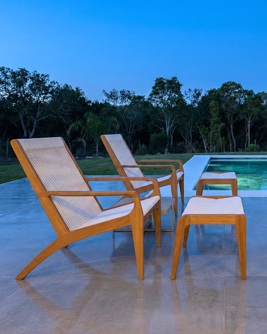 Área de piscina em casa de alto padrão com poltronas de madeira e integração com paisagem natural ao entardecer