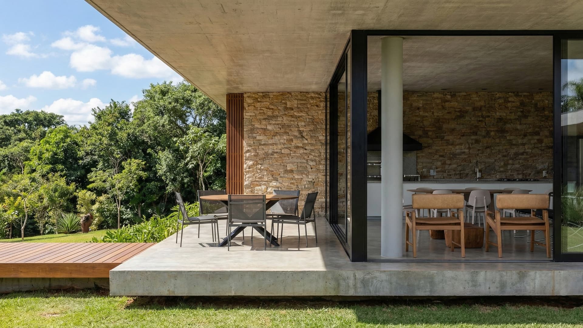 Varanda gourmet integrada ao jardim com laje em balanço de concreto aparente, deck de madeira e parede de pedra natural.