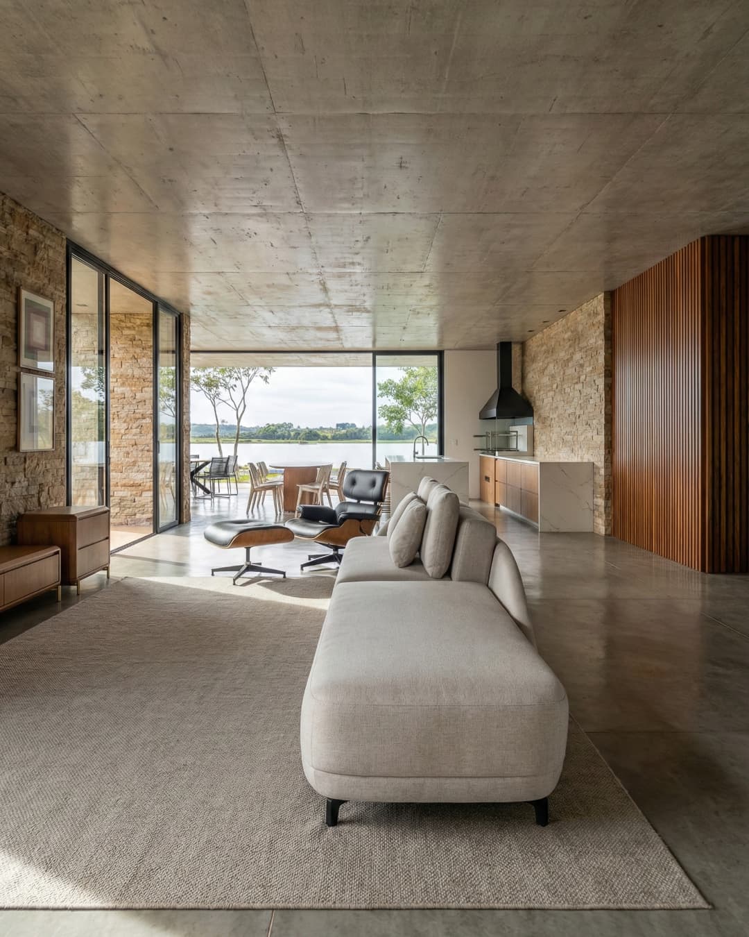 Sala de estar integrada com teto de concreto aparente, parede de pedra natural, poltrona Eames e vista para o lago em casa de luxo.