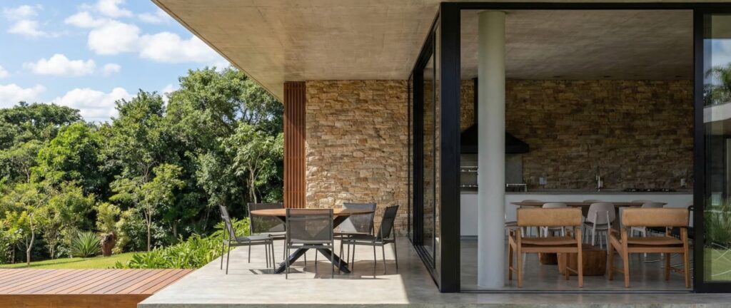 Varanda gourmet de luxo com churrasqueira integrada, parede em pedra natural, forro de concreto aparente, mesa de jantar redonda e mobiliário contemporâneo com vista para jardim arborizado, projetada por arquiteto de alto padrão.