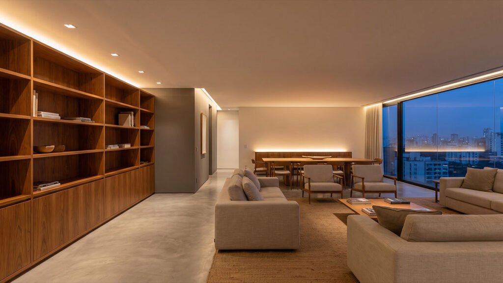 Living integrado de apartamento com marcenaria em madeira, piso cimentício, iluminação indireta e vista da cidade ao entardecer.
