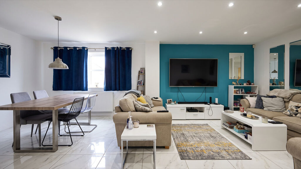 Sala de estar e jantar integradas com parede de destaque azul, painel de TV, piso porcelanato marmorizado e cortinas azuis.