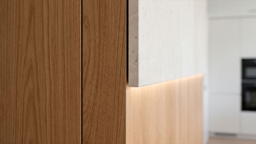 Detalhe do encontro entre painel de madeira e pedra natural clara, com fita de LED embutida iluminando a marcenaria.
