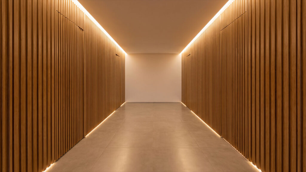 Corredor de alto padrão revestido em painéis ripados de madeira, com iluminação linear embutida no teto e no piso.