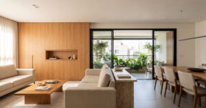 Sala de apartamento com painel de marcenaria em madeira clara, sofás bege, mesa de jantar e varanda envidraçada com plantas